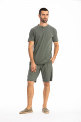 Man wearing a olive green buttoned t-shirt and linen shorts on a white background